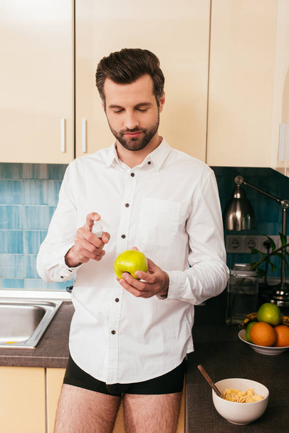 Man in shirt and panties holding apple and hand sanitizer near cereals in kitchen  - 写真・画像