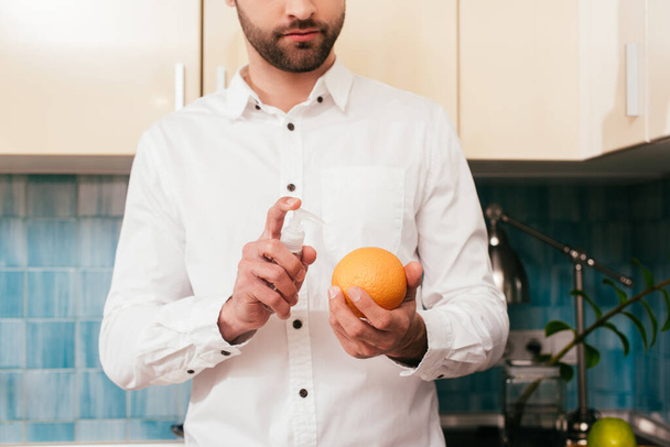 Cropped view of man disinfecting orange with hand sanitizer in kitchen  - Foto, Bild