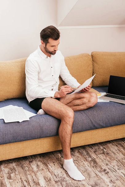 Handsome teleworker in panties and shirt working with papers near laptop on couch  - Foto, afbeelding