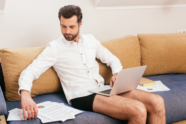 Freelancer in panties and shirt using laptop and working with papers on sofa at home  - Fotó, kép
