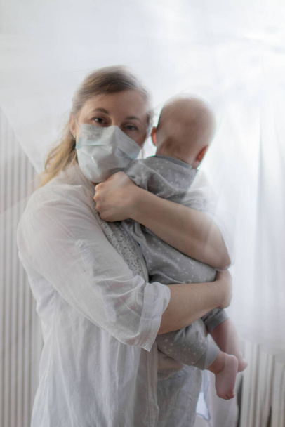 Mother or doctor wearing a face mask holding a baby boy staying inside safe in times of coronavirus pandemic spread in the world near a window - Photo, Image