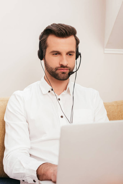 Selective focus of handsome teleworker using laptop and headset at home  - Foto, afbeelding