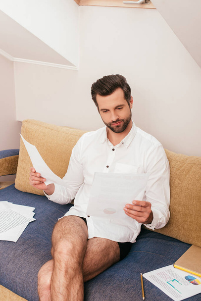 Handsome freelancer in panties and shirt working with papers near book on couch  - Фото, изображение