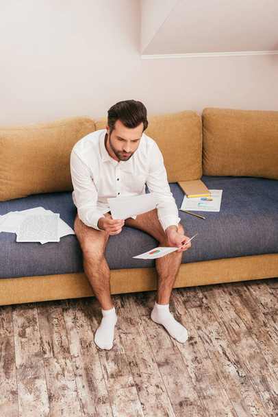 Freelancer in panties and shirt looking at documents with charts on sofa  - 写真・画像