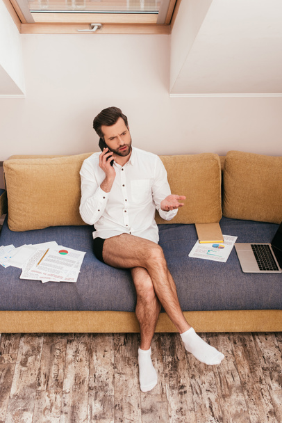 Thoughtful freelancer in panties and shirts taking on smartphone near papers and laptop on couch  - 写真・画像