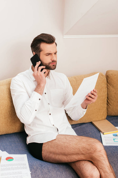 Confident freelancer in shirt and panties talking on smartphone and working with papers on couch   - Fotoğraf, Görsel