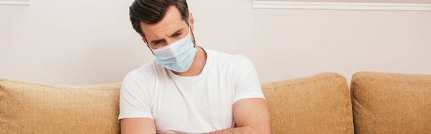 Panoramisch shot van zieke man met medisch masker zittend op de bank thuis  - Foto, afbeelding