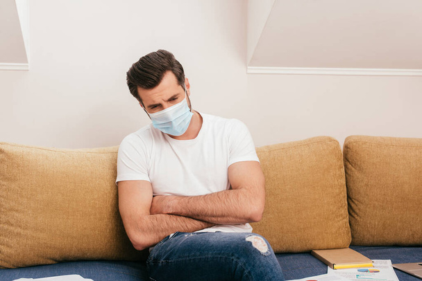 Enfermo freelancer en máscara médica sentado cerca de papeles en el sofá
  - Foto, Imagen