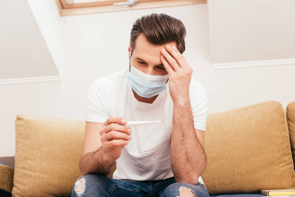 Man in medical mask with hand near forehead holding thermometer on sofa at home  - Photo, Image