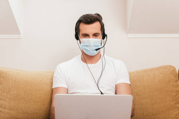 Селективный фокус телеработника в медицинской маске с помощью ноутбука и гарнитуры в гостиной
 - Фото, изображение