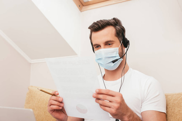 Selective focus of teleworker in medical mask and headset holding document and paper near laptop on couch  - Foto, Imagen