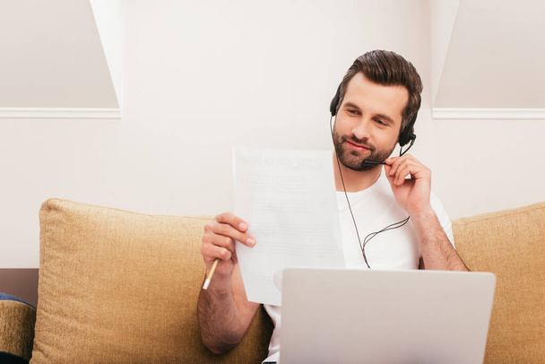 Selective focus of handsome teleworker in headset holding paper near laptop on couch  - Photo, image