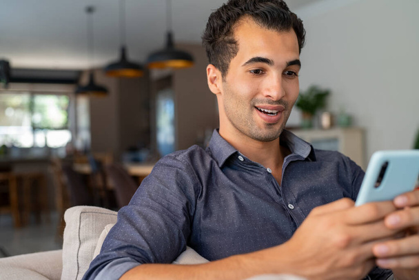 Happy smiling latin man with smartphone being surprised as reading message while sitting on sofa. Indian man sitting on couch looking at his mobile phone with a surprised expression. Hispanic guy with cellphone relaxing at home while receive great ne - Foto, imagen