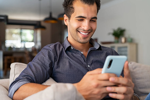 Cheerful businessman using smartphone while sitting on sofa at home. Handsome young indian man sitting on couch reading messages on mobile phone. Hispanic guy working from home with cellphone. - Photo, image