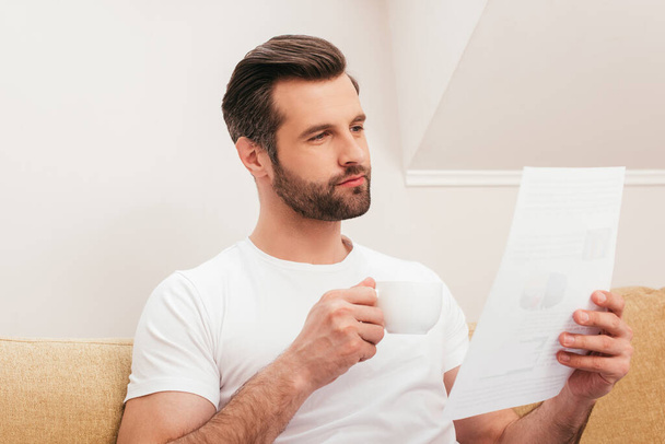 Handsome teleworker holding document and drinking coffee on couch   - Фото, зображення