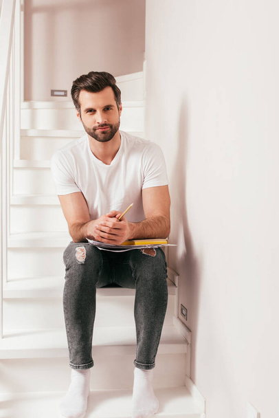 Handsome man holding pencil and book with documents on staircase at home  - Foto, Bild