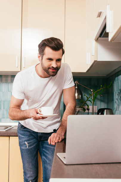 Valikoiva painopiste hymyilevä mies kuppi kahvia katsomalla kannettavaa keittiön työtaso
  - Valokuva, kuva