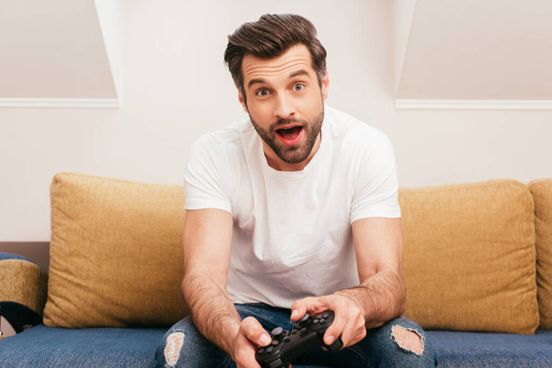 KYIV, UKRAINE - APRIL 14, 2020: Shocked man holding joystick on couch at home  - Foto, immagini