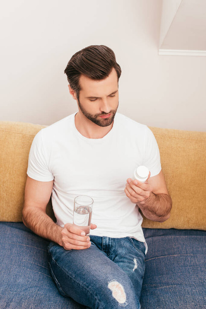 Handsome man holding jar with pills and glass of water on couch at home  - Valokuva, kuva
