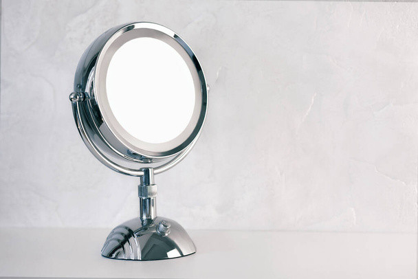 Chromed steel round magnifying mirror on handle set up on the table. Cold light background, the atmosphere of purity and cleanliness. Studio, copy space for any text or design work. - Fotó, kép