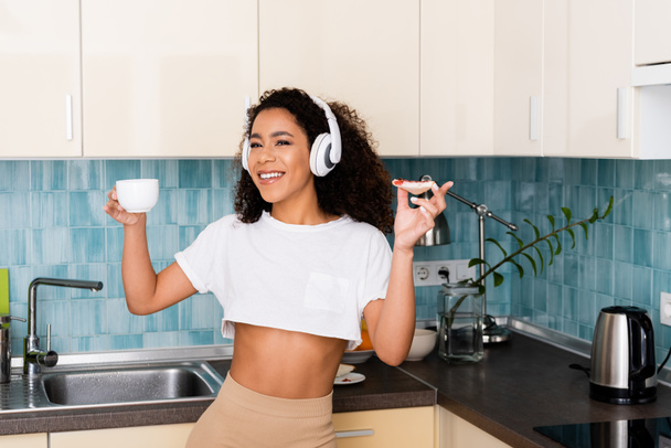 cheerful african american girl in wireless headphones holding cup and toast bread with jam  - Foto, Imagem