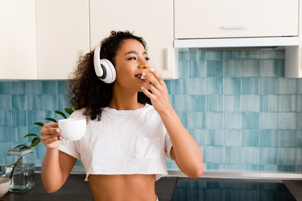 chica afroamericana feliz en auriculares inalámbricos sosteniendo taza y comer pan tostado con mermelada
 - Foto, Imagen