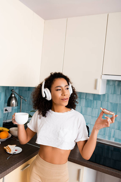curly african american girl in wireless headphones holding toast bread with jam and cup of coffee  - Foto, Bild