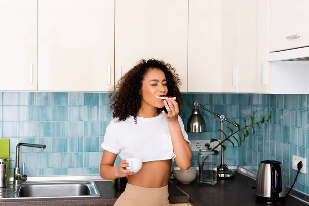 χαρούμενο αφροαμερικανό κορίτσι τρώει ψωμί τοστ με μαρμελάδα και κρατώντας ένα φλιτζάνι καφέ  - Φωτογραφία, εικόνα