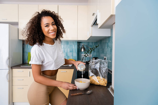 happy african american woman pouring milk in bowl with cornflakes  - Foto, imagen