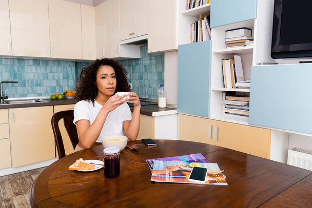 魅力的なアフリカ系アメリカ人の女性がおいしい朝食の近くにコーヒーを持ってカップを保持し、空白の画面を持つスマートフォン  - 写真・画像