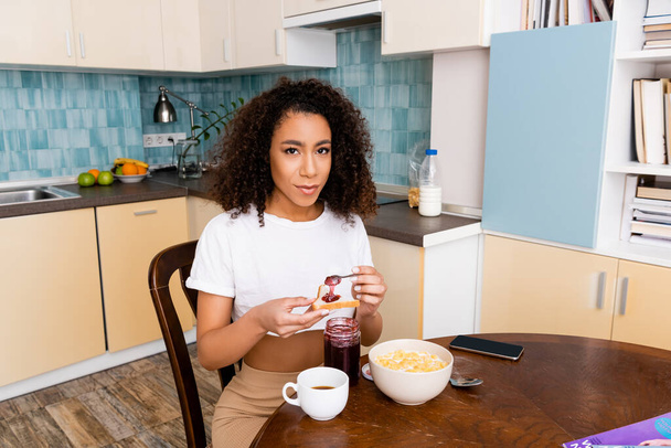 curly african american woman holding spoon with sweet jam near toast bread and smartphone with blank screen  - Foto, Bild