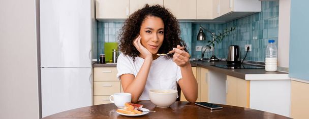 raccolto panoramico di donna afro-americana che tiene cucchiaio con corn flakes vicino a tazza con caffè e smartphone con schermo bianco sul tavolo
  - Foto, immagini