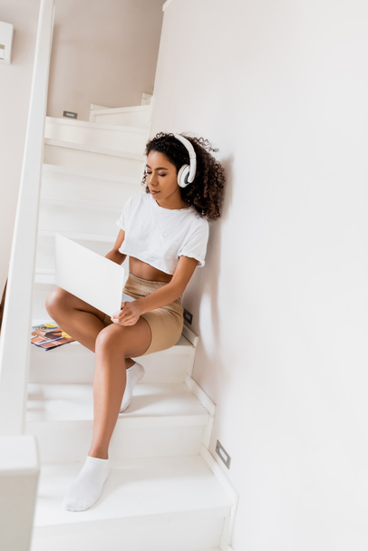 афроамериканський фрілансер в бездротових навушниках, що сидять на сходах і використовують ноутбук вдома
 - Фото, зображення