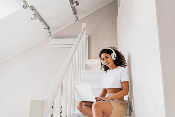 beautiful african american freelancer in wireless headphones sitting on stairs and using laptop  - Фото, изображение