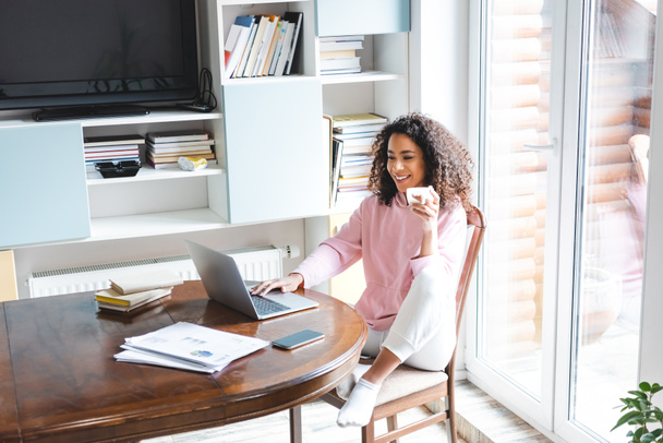 happy african american freelancer holding cup while using laptop at home  - Photo, Image