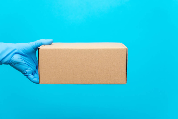 Un hombre sostiene una caja de cartón con la mano en un guante de goma. Protección contra COVID-19. Entrega segura de bienes durante la epidemia de coronavirus
. - Foto, Imagen