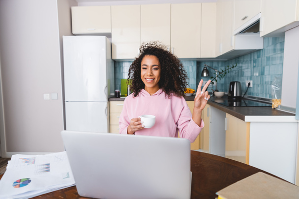 happy african american girl showing peace sign and holding cup while having video chat at home  - Φωτογραφία, εικόνα