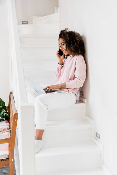 cheerful african american freelancer sitting on stairs while using laptop and talking on smartphone  - Photo, Image