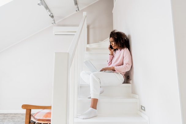 displeased african american freelancer sitting on stairs while using laptop and talking on smartphone  - Fotografie, Obrázek