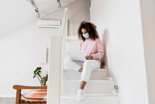 young african american freelancer in medical mask sitting on stairs and using laptop  - Fotó, kép
