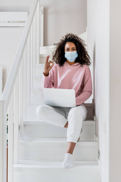 curly african american freelancer in medical mask sitting on stairs and showing ok sign near laptop  - Valokuva, kuva