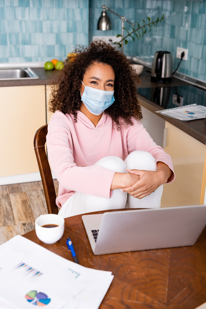 curly african american freelancer in medical mask sitting near laptop, cup, charts and graphs  - Φωτογραφία, εικόνα