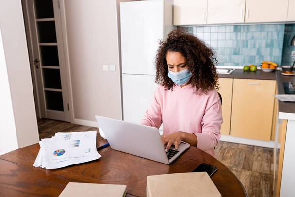 young african american freelancer in medical mask using laptop near cup, charts and graphs  - Foto, Bild