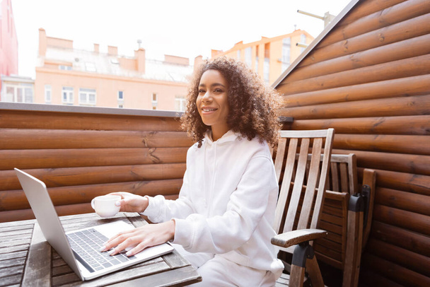 happy african american freelancer holding cup and using laptop outside  - Photo, Image
