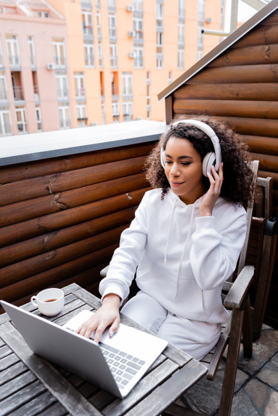 curly african american freelancer touching wireless headphones and using laptop near cup  - Photo, Image