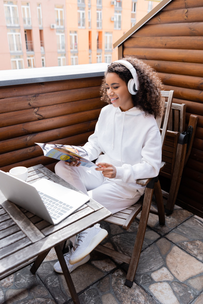 boldog afro-amerikai szabadúszó vezeték nélküli fejhallgató olvasás magazin közelében laptop és csésze  - Fotó, kép