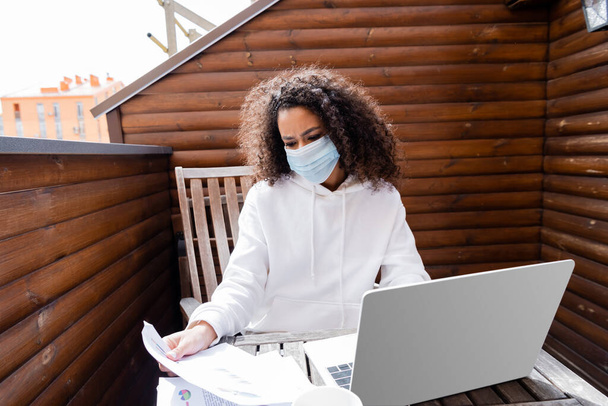 african american girl in medical mask looking at charts and graphs near laptop - Zdjęcie, obraz
