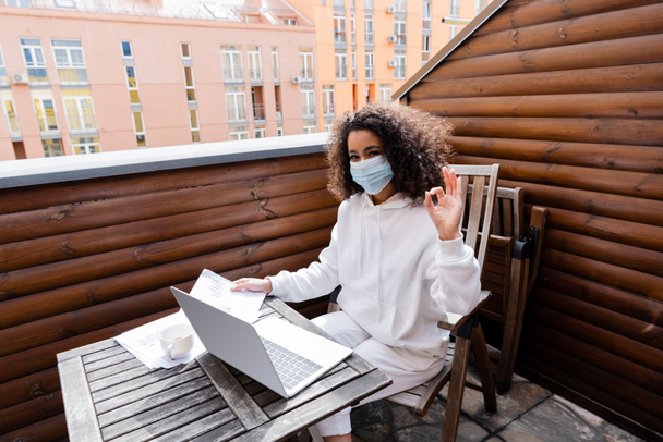 afro-amerikai lány orvosi maszk mutatja rendben jel közelében laptop, csésze, diagramok és grafikonok  - Fotó, kép