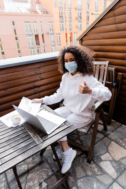 african american girl in medical mask showing thumb up near laptop, charts and graphs  - 写真・画像