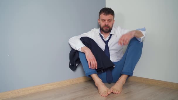 Uomo barbuto sconvolto in abiti formali disordinati è seduto sul pavimento in un appartamento vuoto. Gli effetti del virus Covid-19 sull'economia sono la disoccupazione, la povertà e le malattie
. - Filmati, video